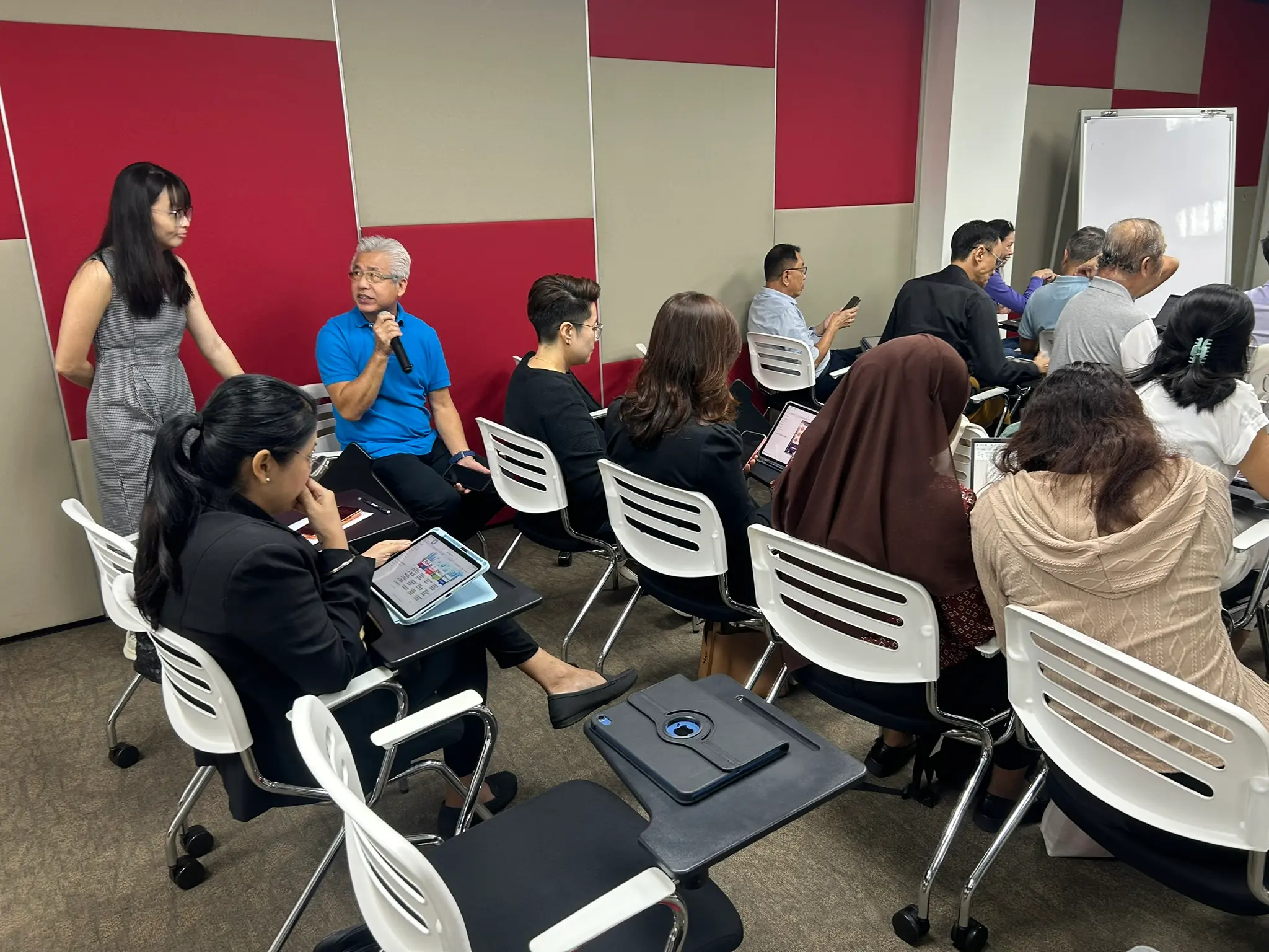 An advisor in a blue shirt speaking into a microphone while seated among the group, with a trainer standing nearby, Penang, 21 September 2026