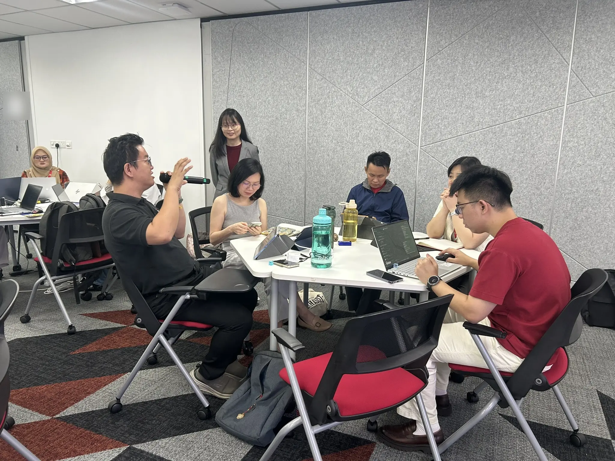 An advisor speaking into a microphone at his table while the trainer stands behind, smiling, Johor Bahru, 14 October 2026