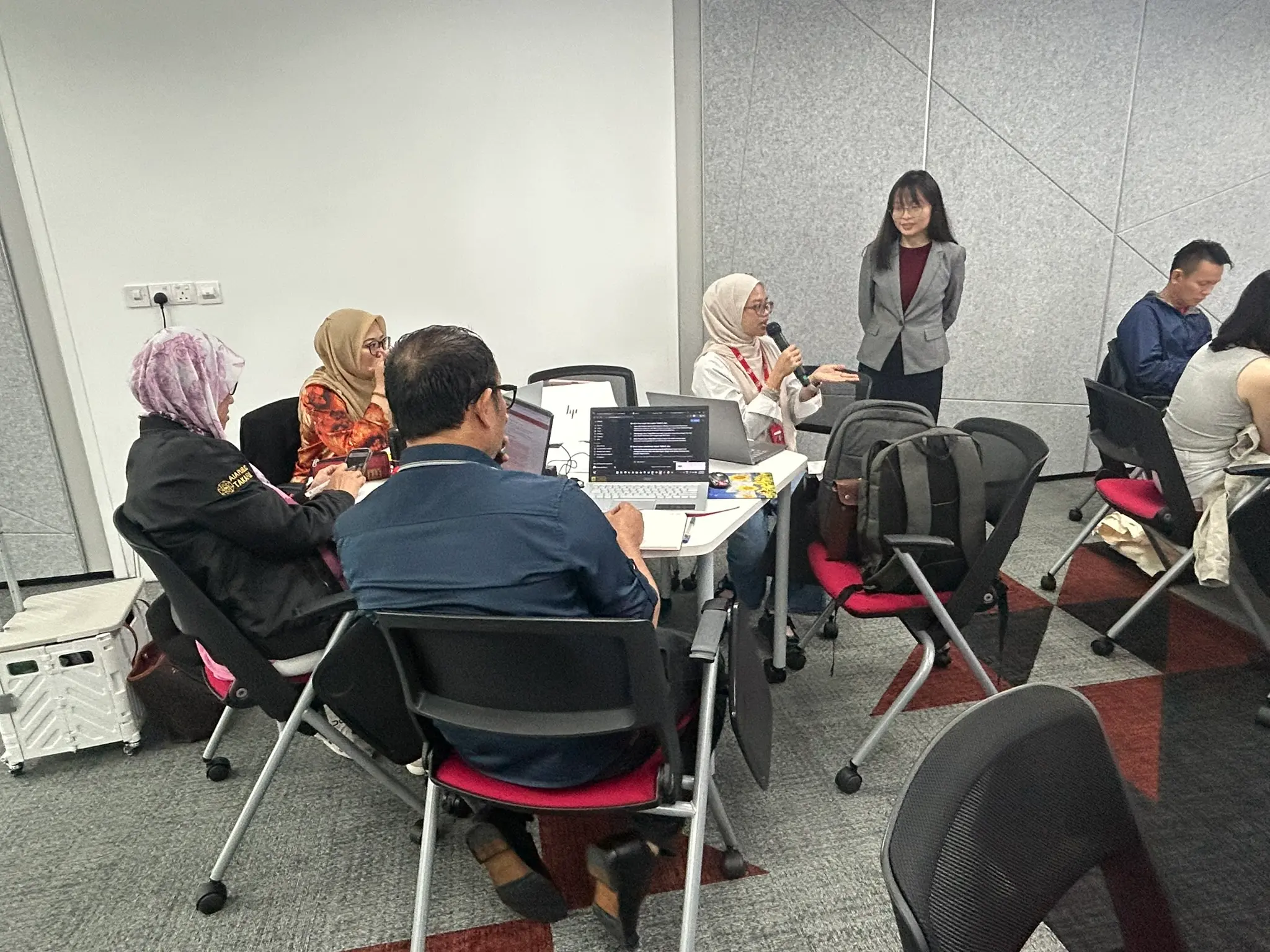 An advisor holding a microphone explains her work to her table as the trainer listens, Johor Bahru, 14 October 2026