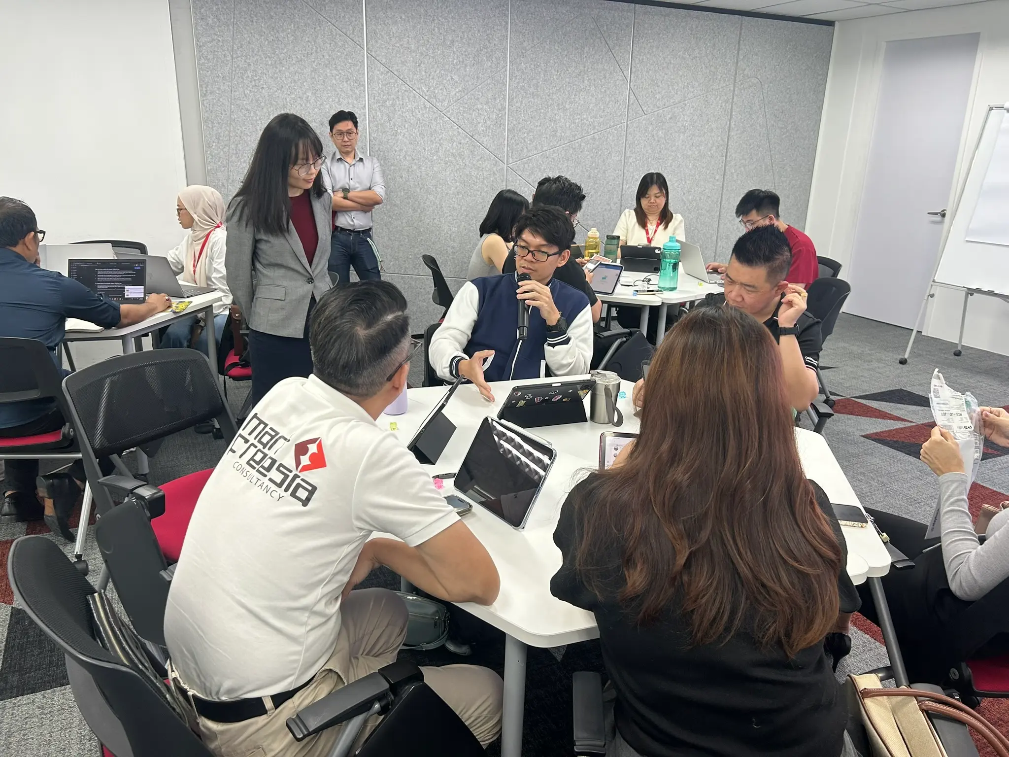 The trainer standing beside a table of advisors working on tablets and laptops while one speaks into a microphone, Johor Bahru, 14 October 2026