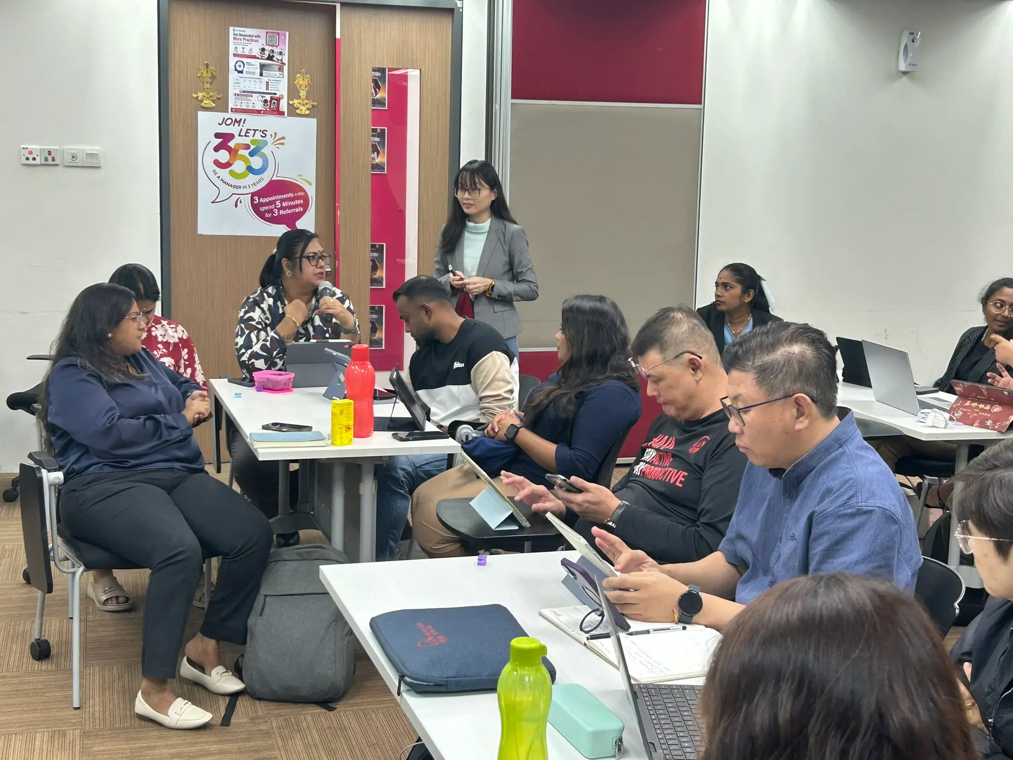 An advisor speaking into a microphone at a table of colleagues with tablets and laptops, while a trainer stands and listens, Seremban, 8 October 2026