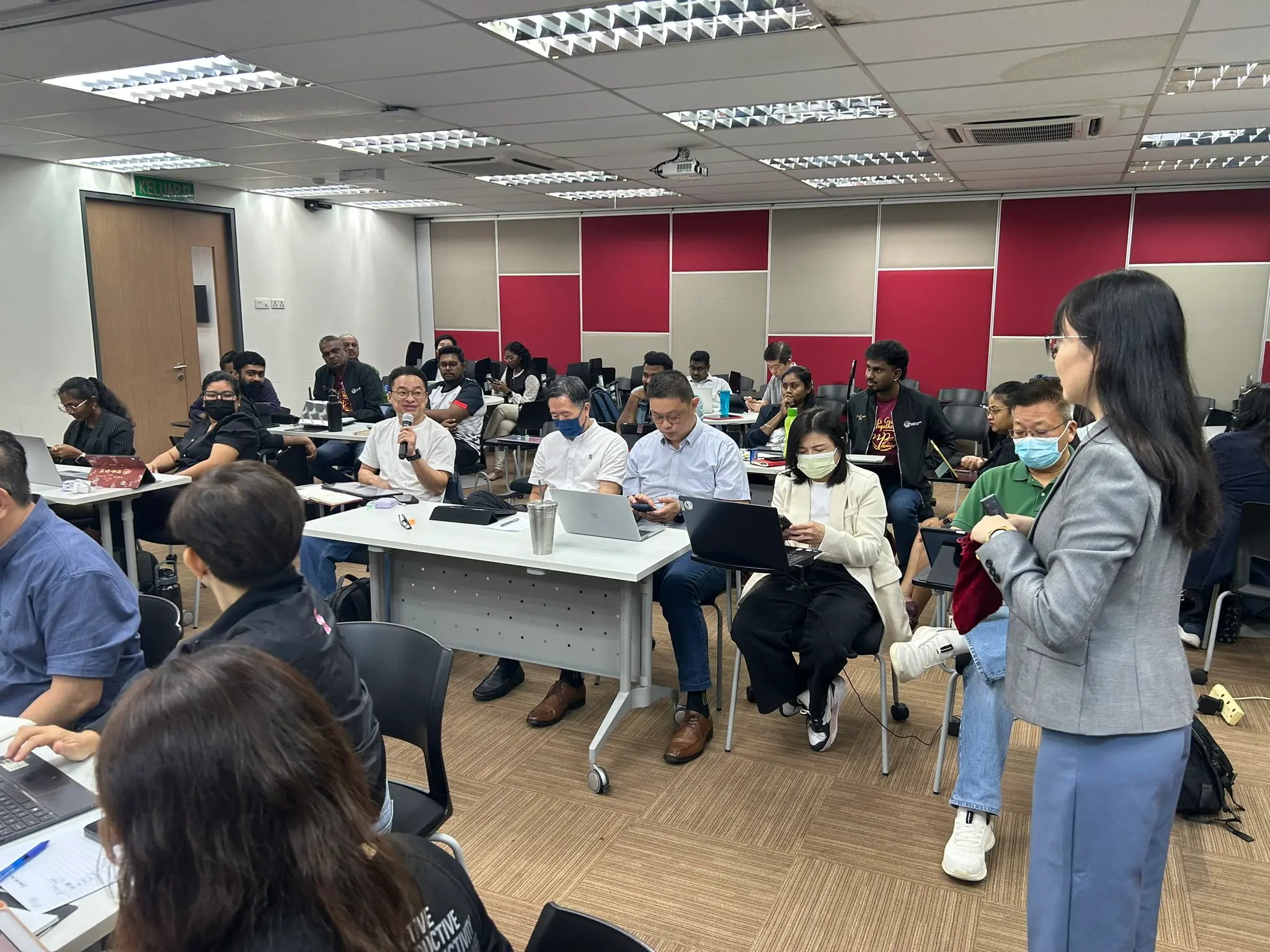 A smiling advisor holding a microphone while seated among rows of colleagues with laptops, as a trainer stands at the side of the room, Seremban, 8 October 2026