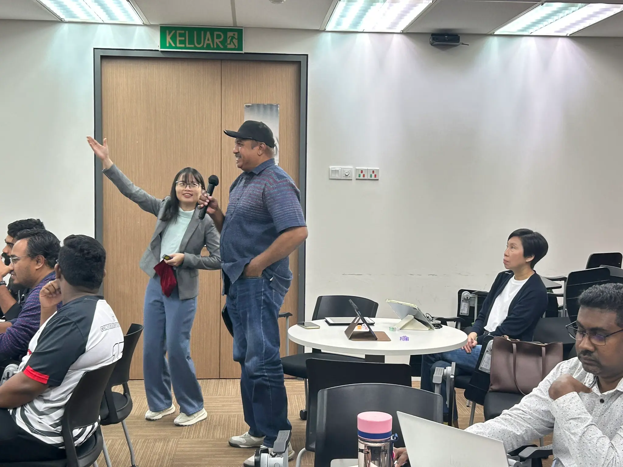 A trainer gesturing beside an advisor in a cap who is speaking into a microphone, with other advisors seated nearby, Seremban, 8 October 2026