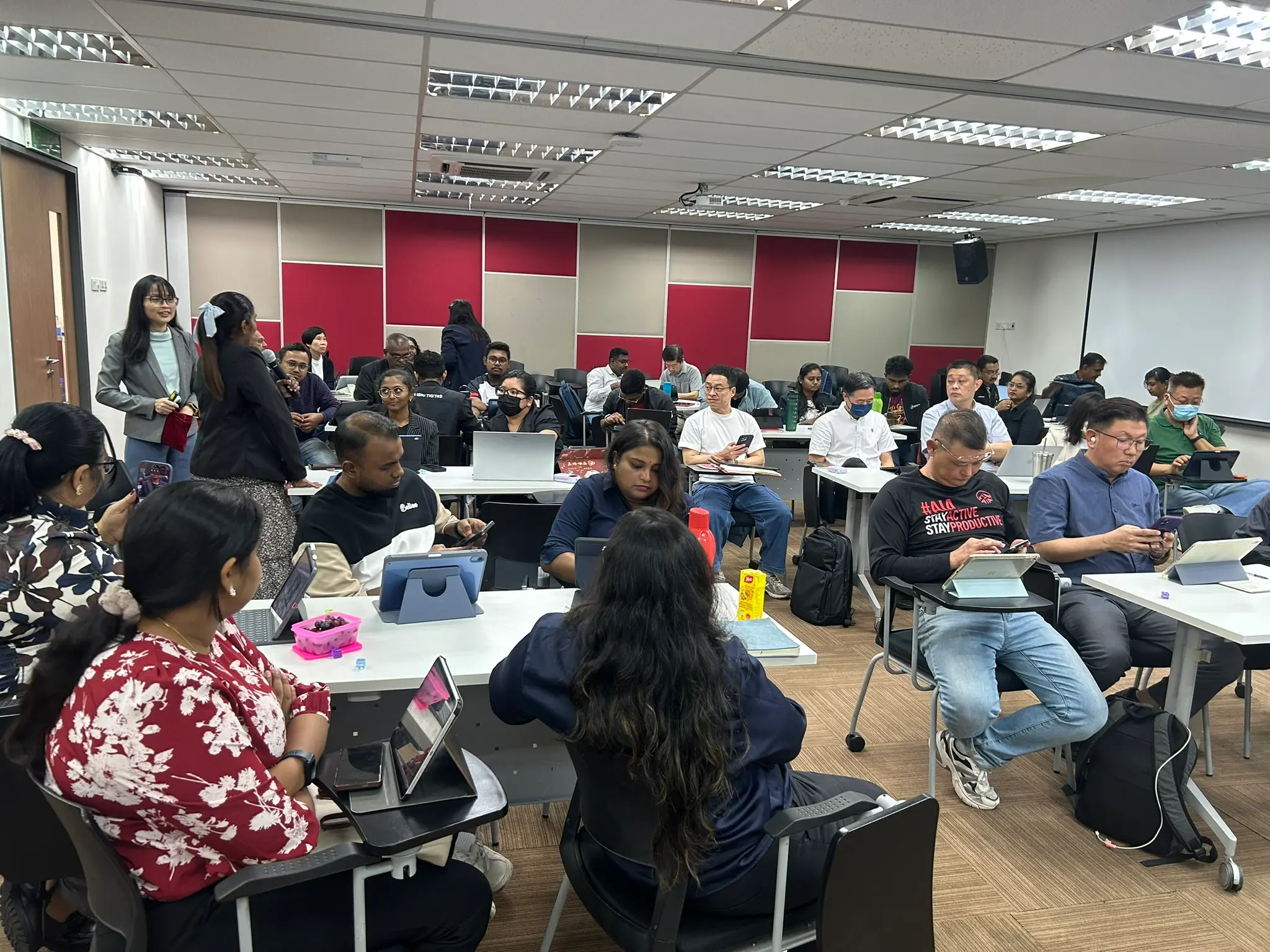 A full training room of advisors working on tablets and phones at long tables, with a trainer and an advisor holding a microphone standing at the side, Seremban, 8 October 2026