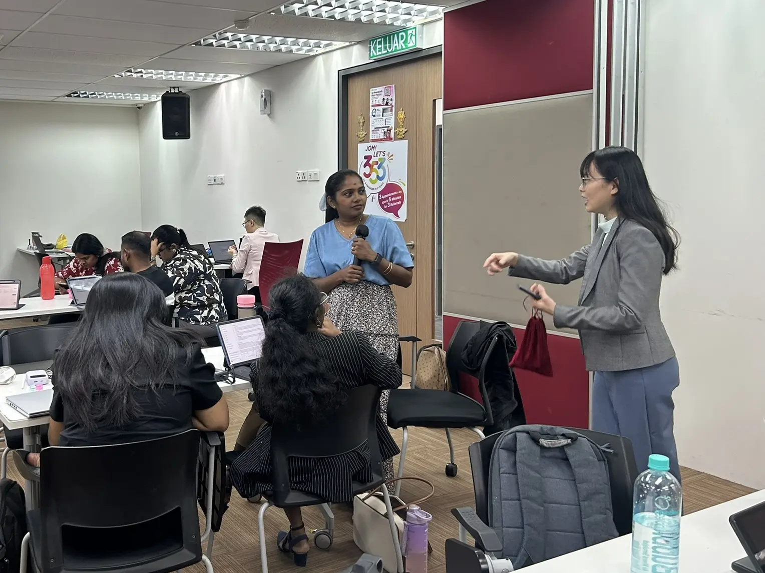 An advisor holding a microphone answering a question from a trainer, while others work on laptops behind them, Seremban, 8 October 2026