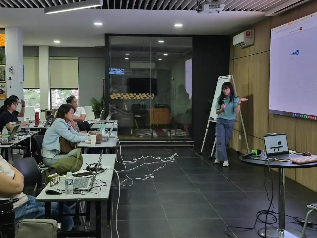 The trainer holding a microphone and explaining a point beside a flip chart while participants listen at their laptops, Kota Kinabalu, 30 September 2026