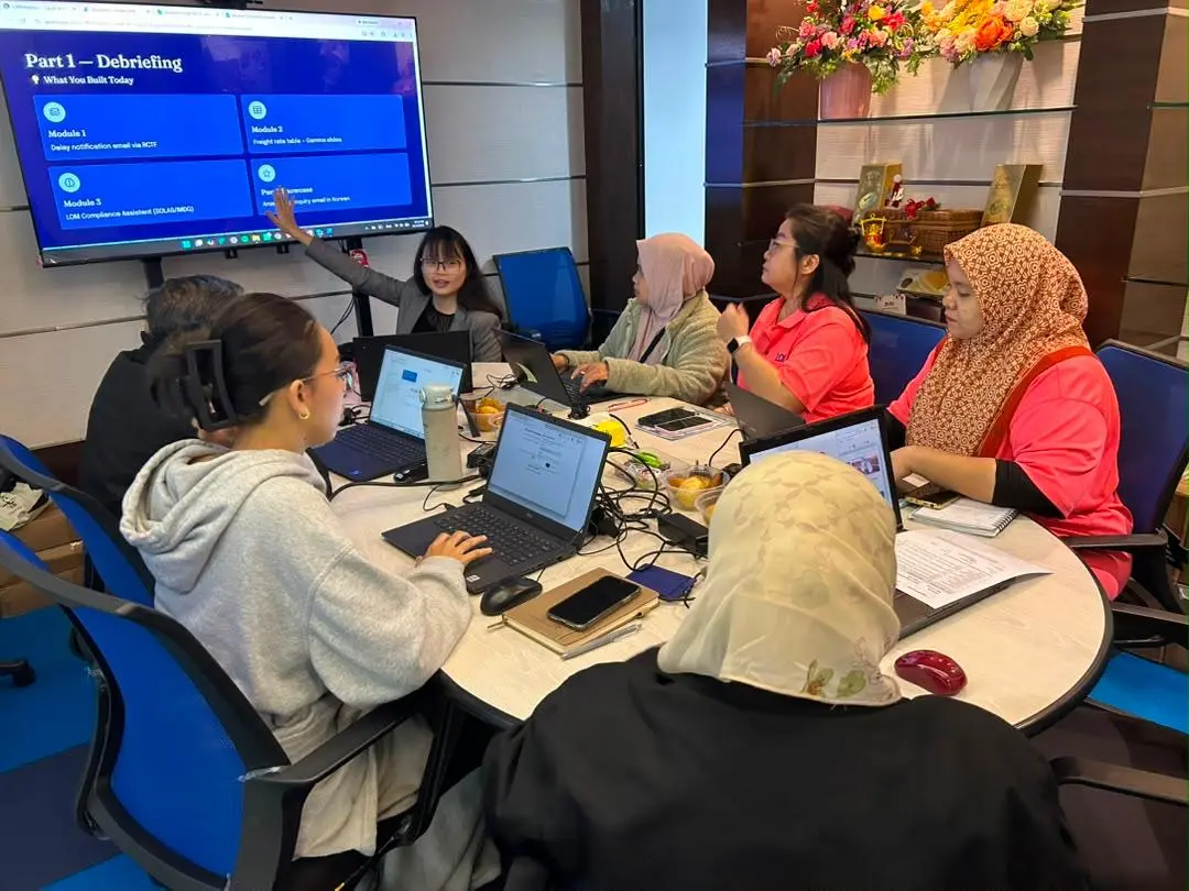 Trainer pointing at a debriefing slide headed What You Built Today, listing the modules completed, with participants at laptops around the table