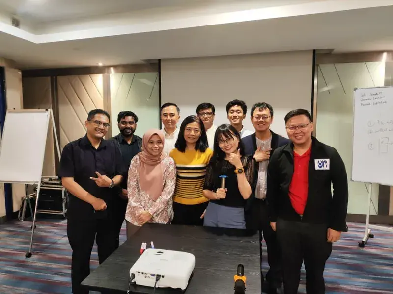 A group of participants with the trainer at the end of an AICoach.my classroom session, whiteboard and projector visible