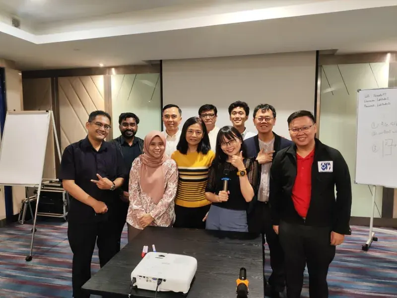 Group photo of participants and trainers at the AI Builder for Work session, one holding the presenter remote, the whiteboard still covered in the day's working notes