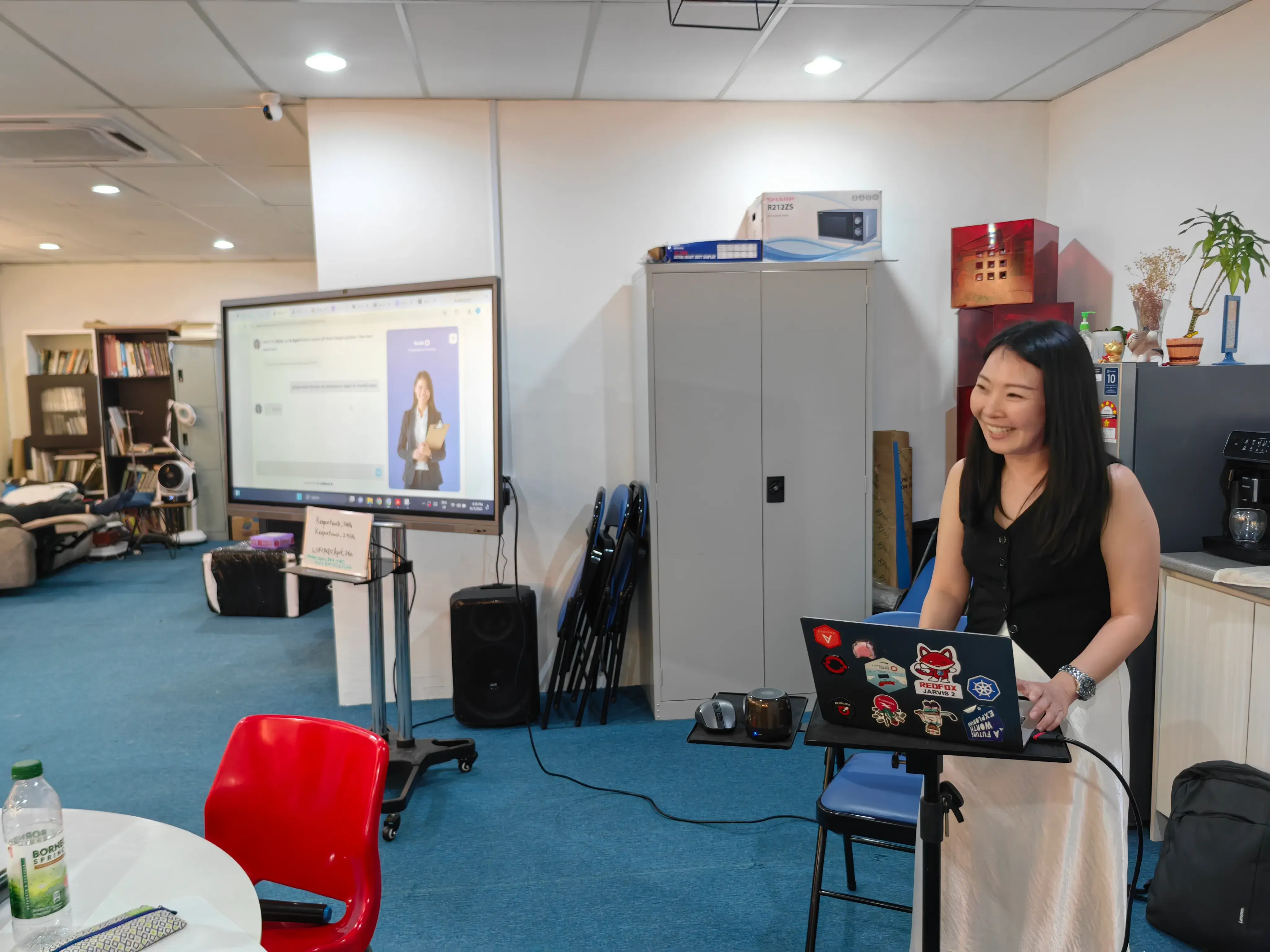 Speaker at a standing laptop with an AI assistant demo on the screen behind her at Inno Day #1