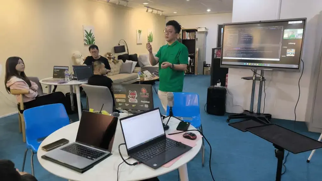 Speaker in a green polo presenting with code and a video call on the screen behind him at Inno Day #2