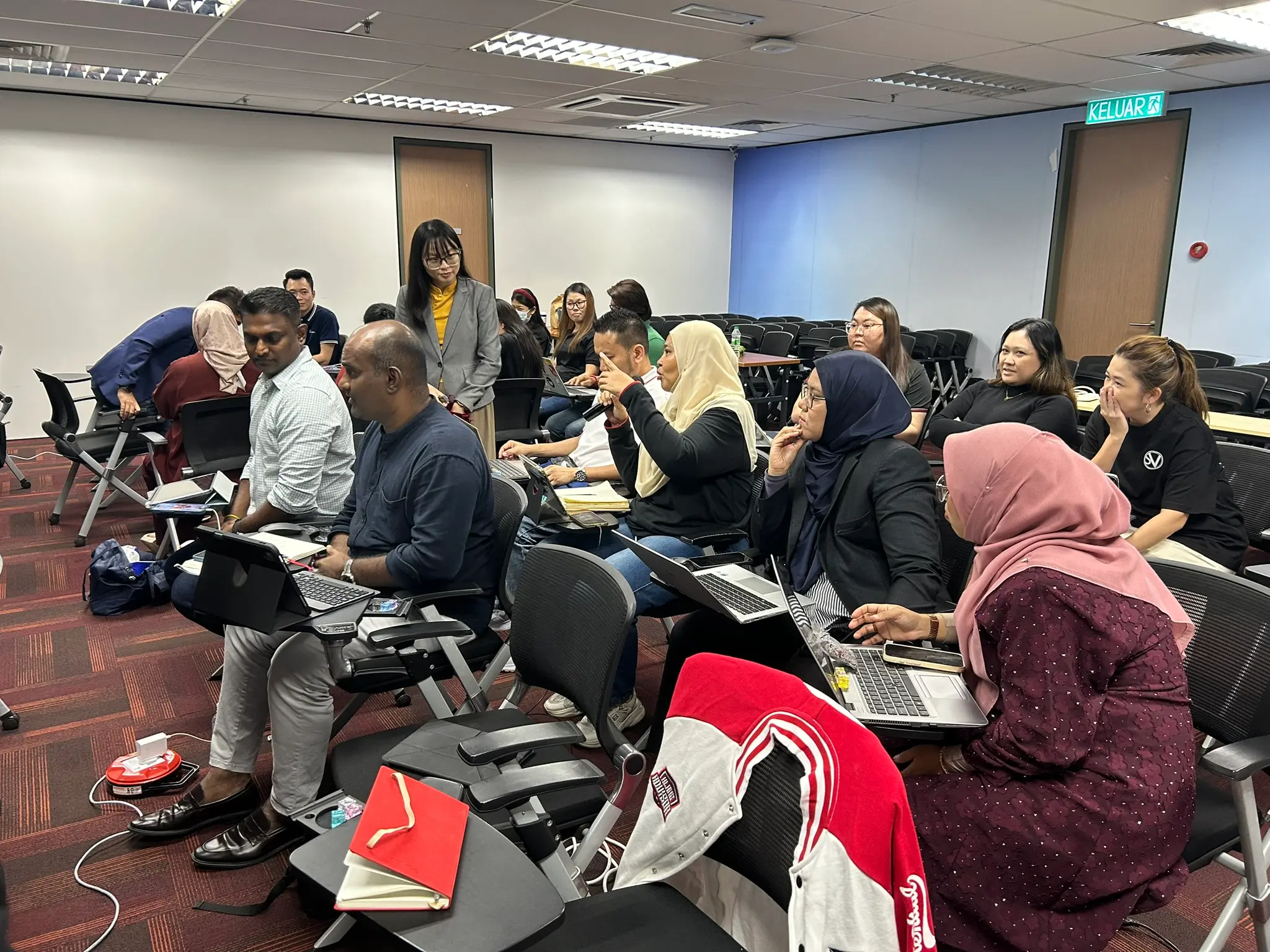 An advisor speaking into a microphone and gesturing while colleagues around her listen and smile, with a trainer standing behind them, Klang, 9 October 2026