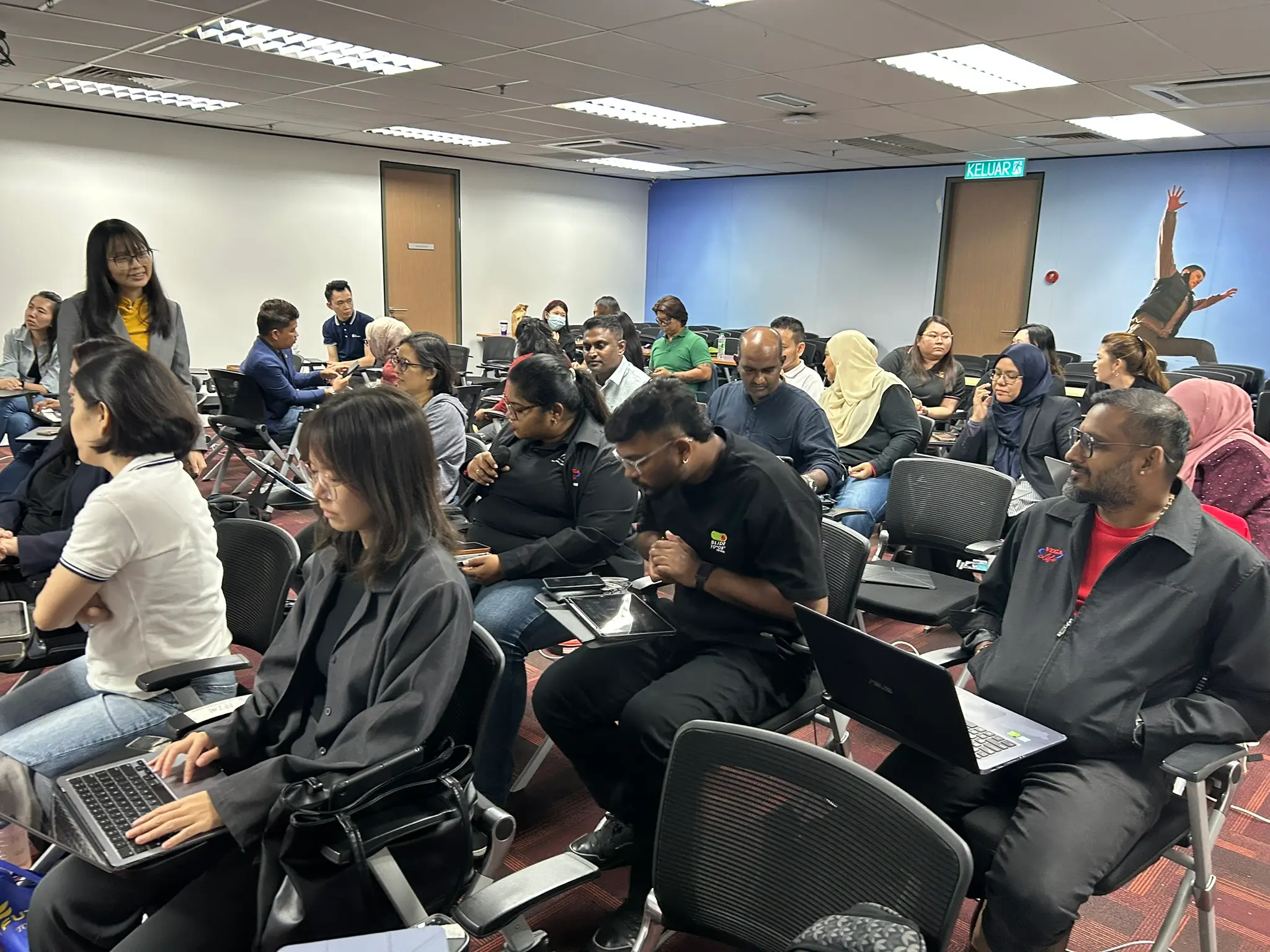 A smiling trainer standing among seated advisors working on laptops and tablets, while one advisor holds a microphone, Klang, 9 October 2026