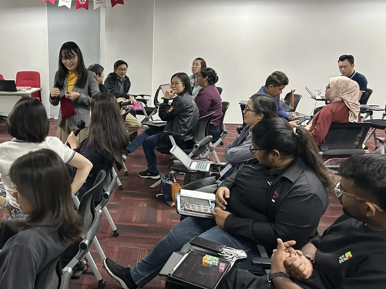 A trainer smiling as she talks with advisors seated in a loose circle of chairs, one of them holding a microphone, Klang, 9 October 2026