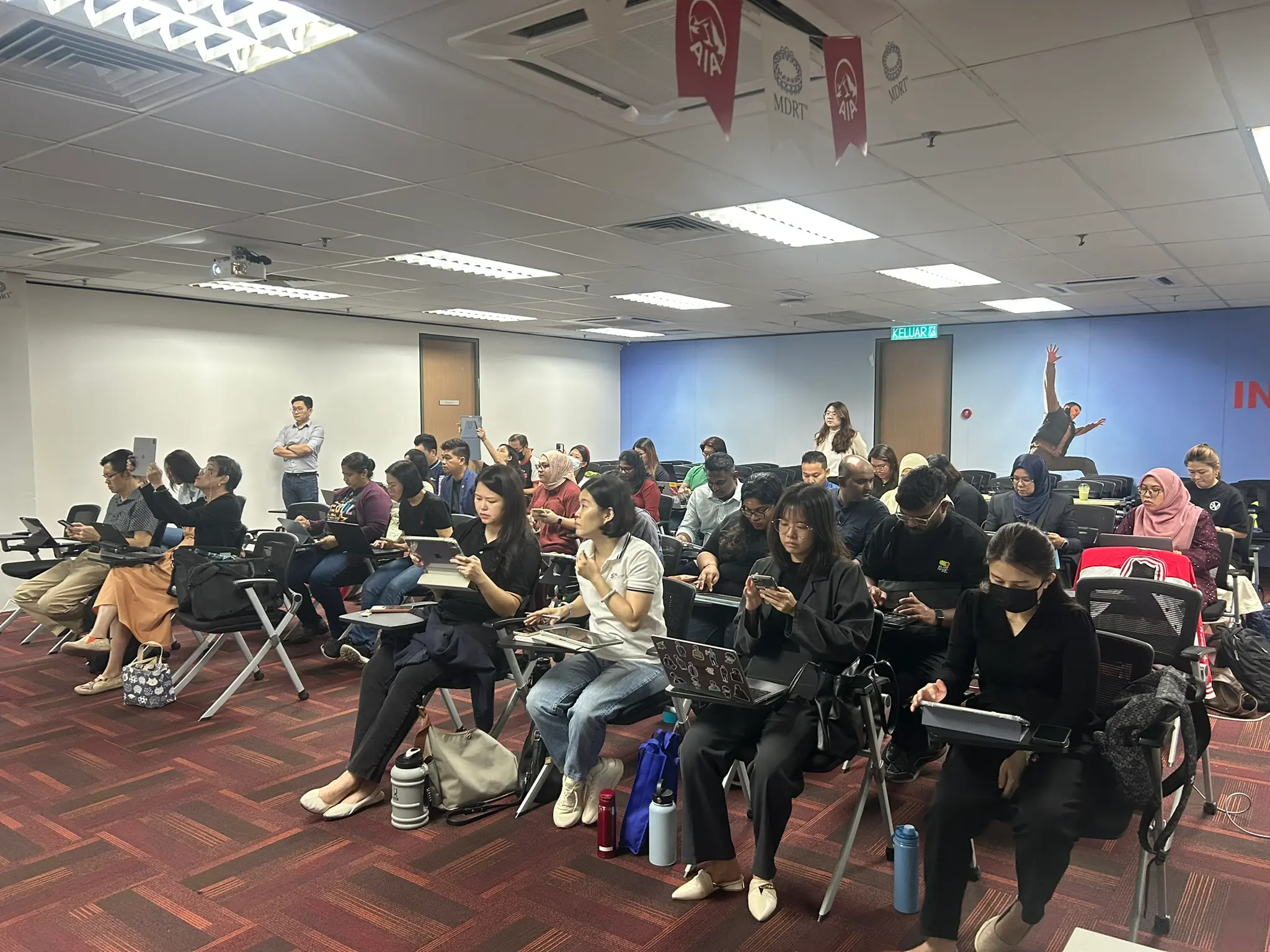 Rows of advisors working on tablets, laptops and phones in a training room with a blue wall mural, Klang, 9 October 2026