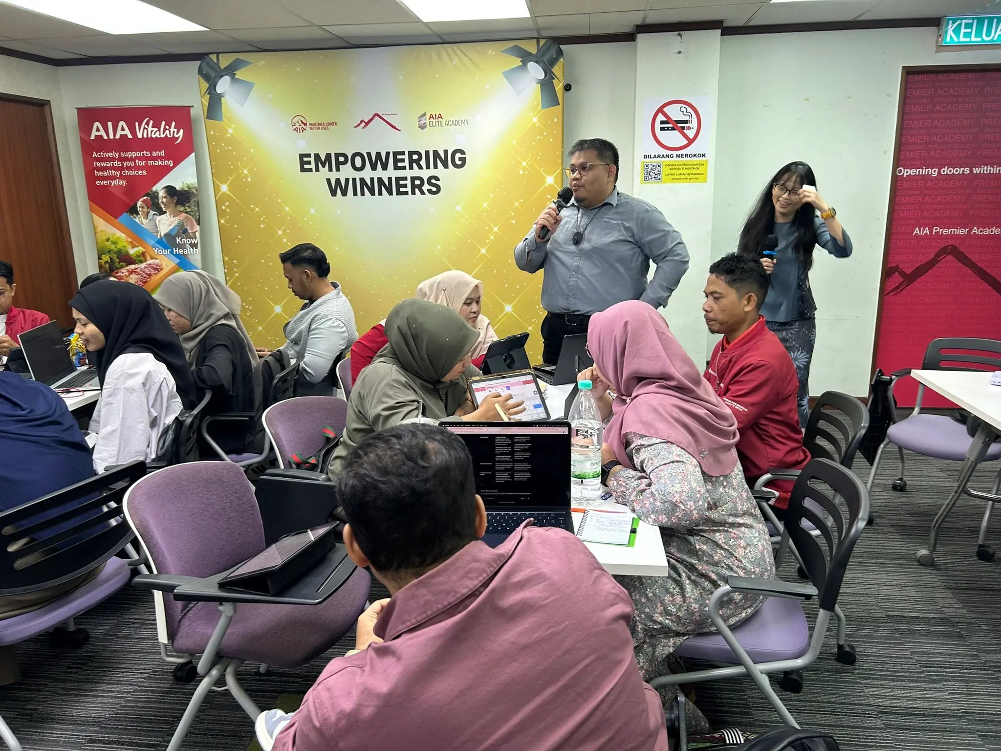 Seorang peserta berdiri berkongsi dengan mikrofon, jurulatih mendengar di sebelah