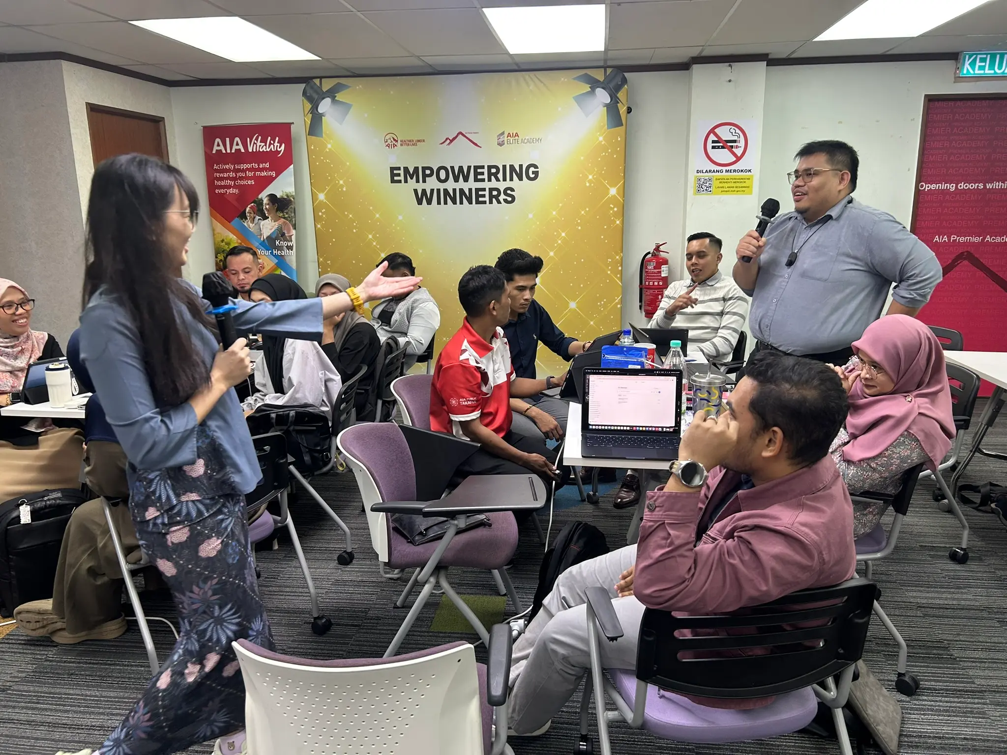 Jurulatih menghulurkan tangan ke arah peserta yang sedang bercakap, peserta lain tersenyum