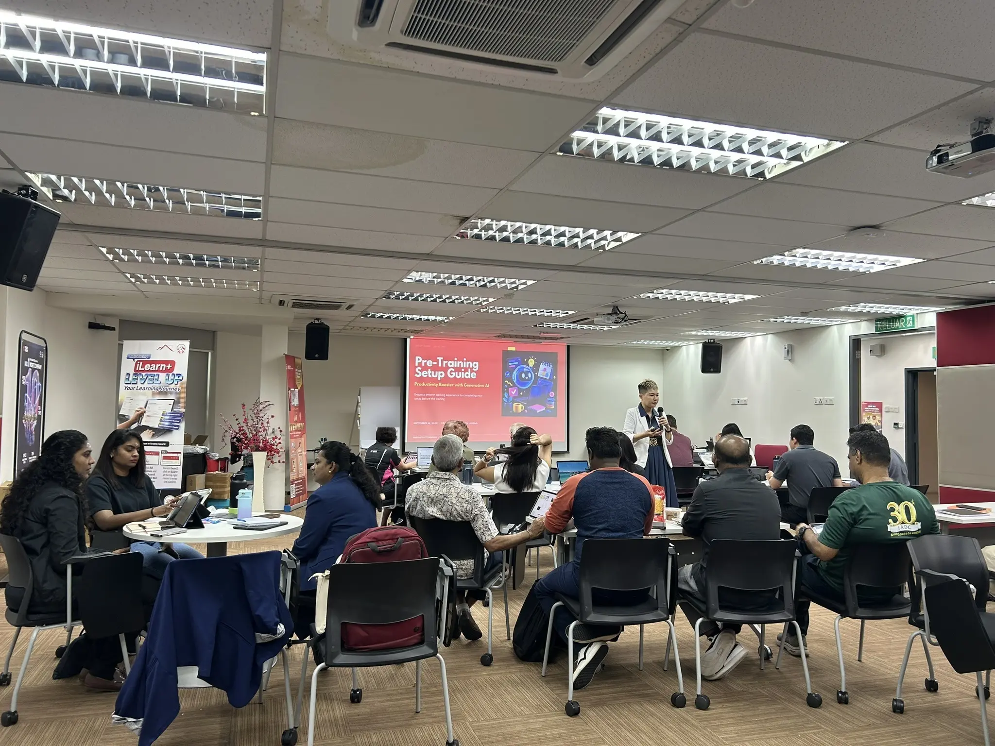A trainer speaking into a microphone at the front of a training room while advisors work on laptops and tablets at round tables, Seremban, 18 September 2026