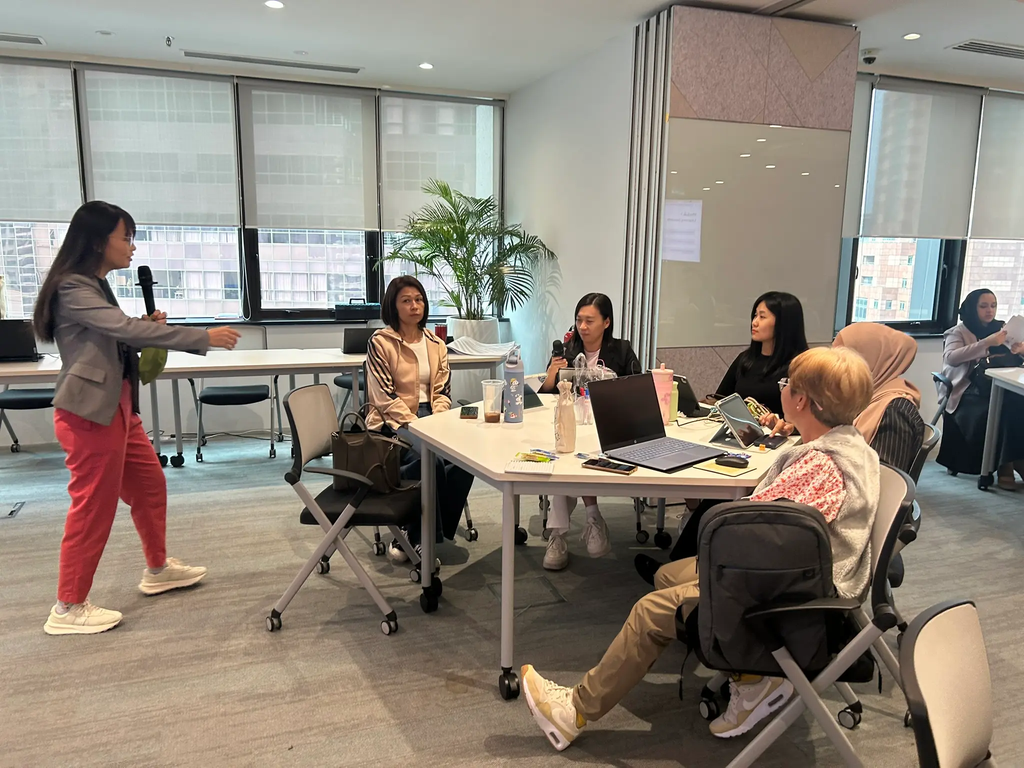 The trainer standing with a microphone beside one small table of advisors