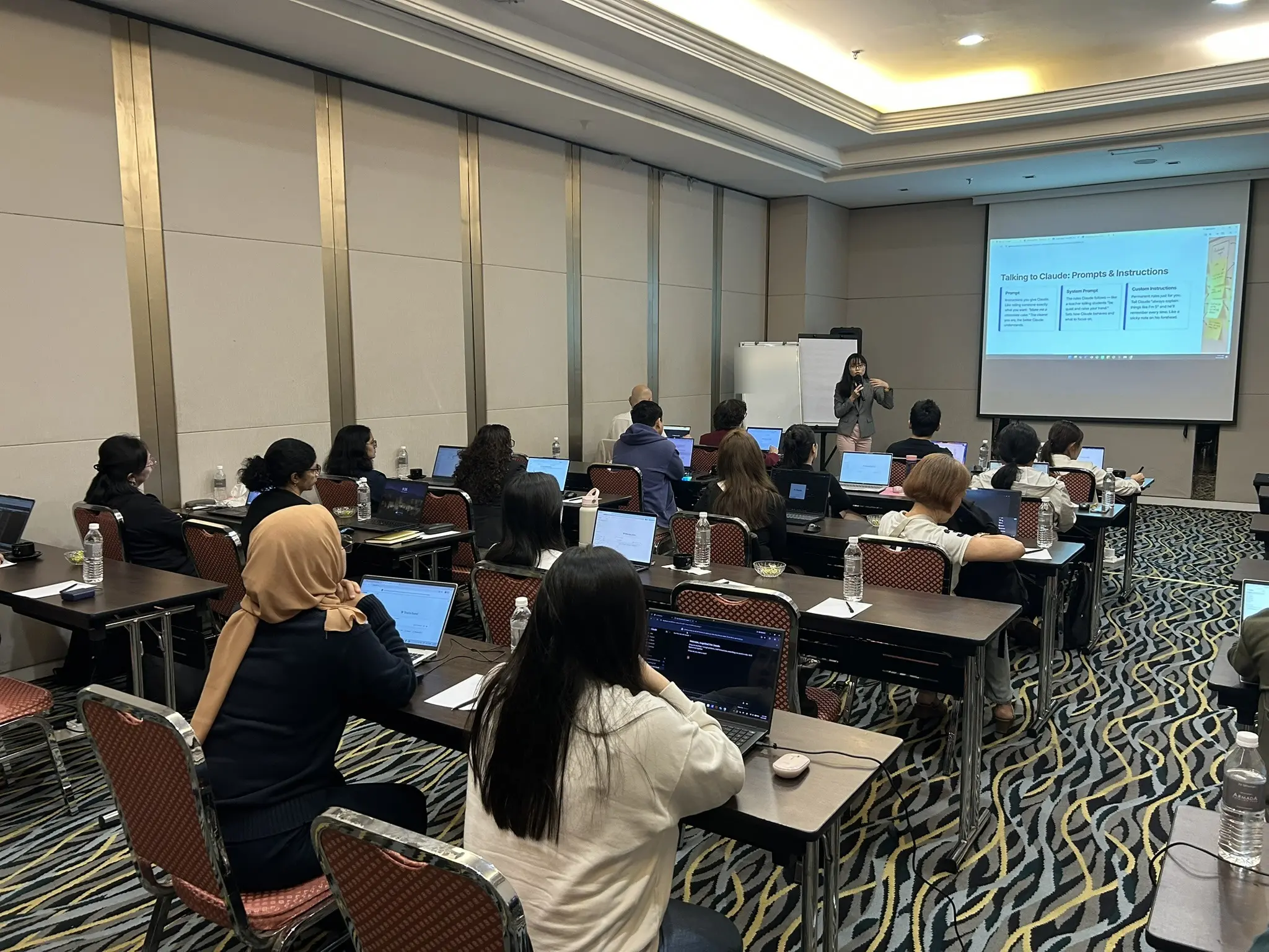 A trainer with a microphone explaining a slide on prompts and instructions to participants working on laptops, Armada Hotel, Petaling Jaya, 24 September 2026