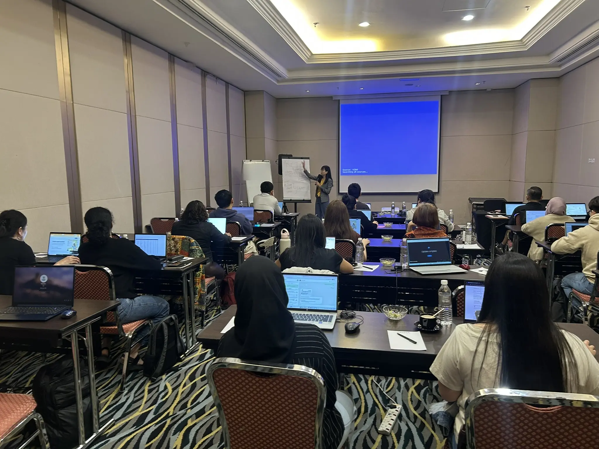 A trainer writing on a flip chart at the front of a hotel function room while participants follow along on their laptops, Petaling Jaya, 25 September 2026