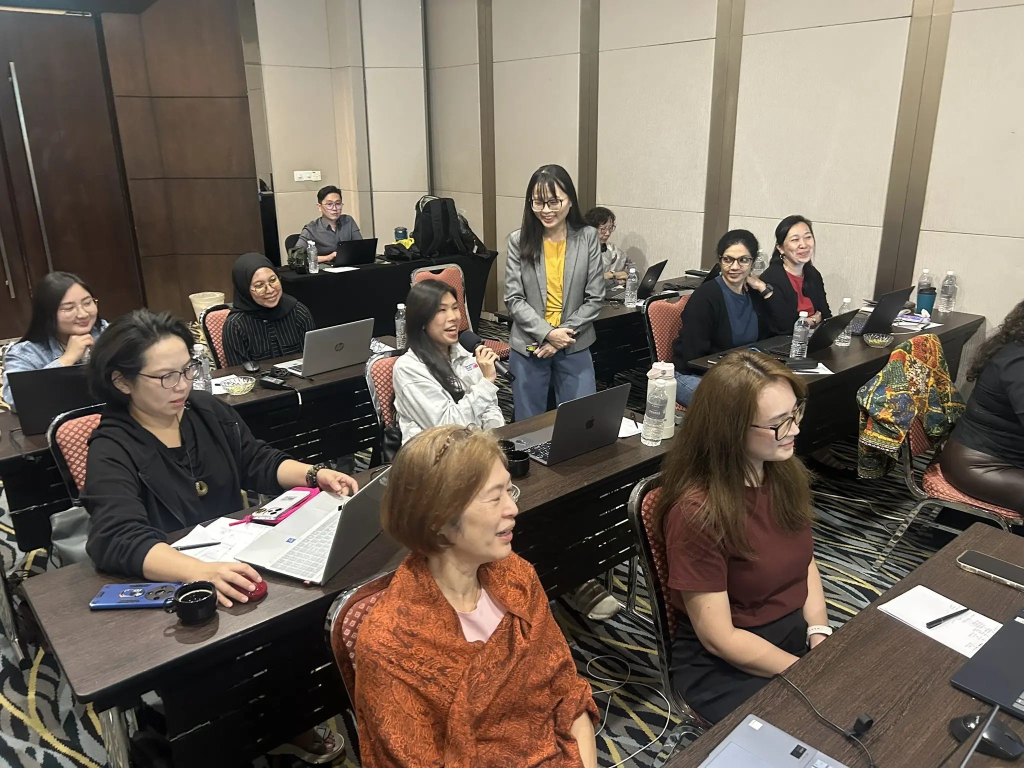 A participant speaking into a microphone while the trainer and others at nearby tables smile and listen, Petaling Jaya, 25 September 2026